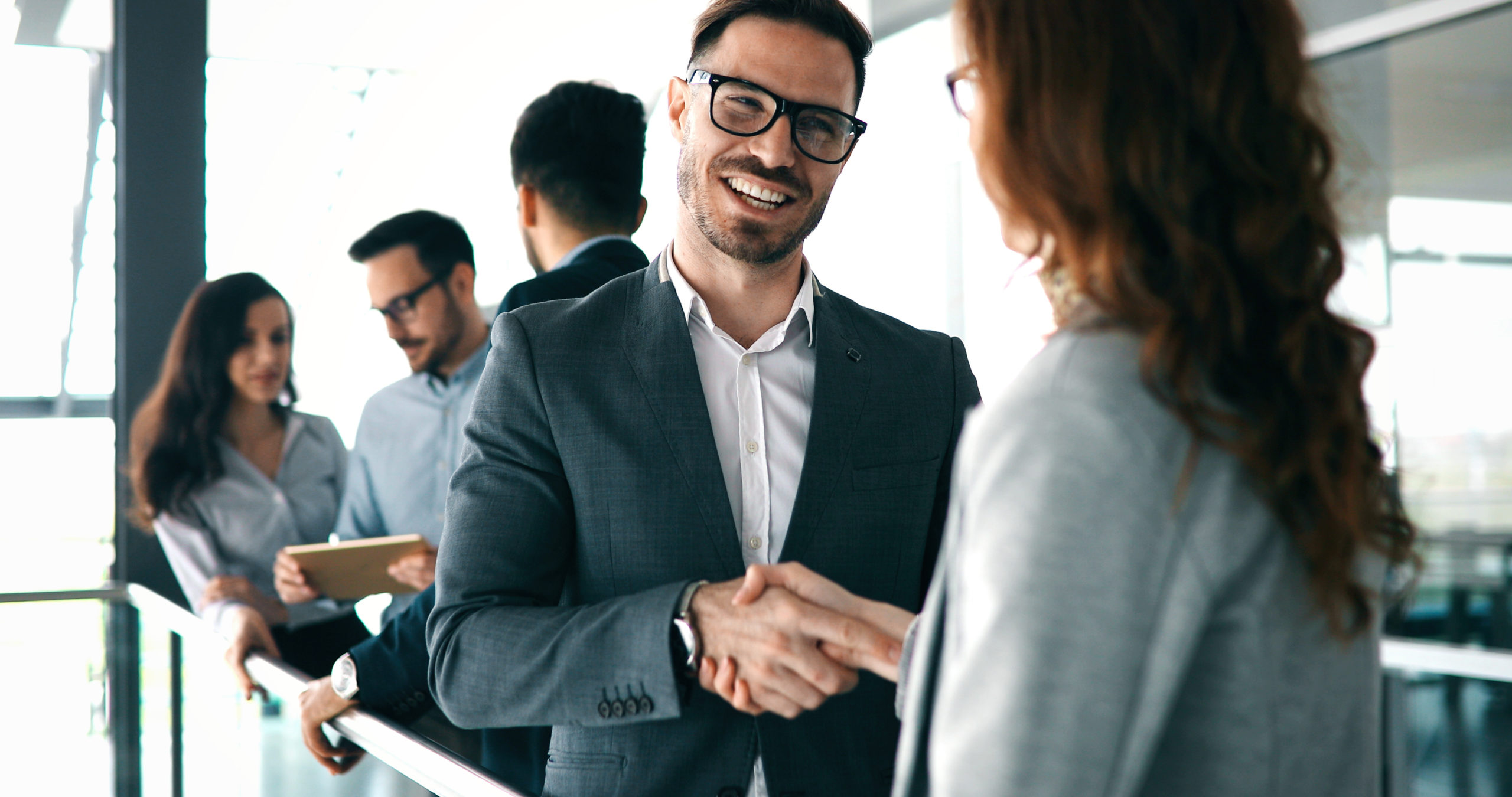 Male consultant shaking hands with female client standing in an office setting with engaged employees talking together in the background