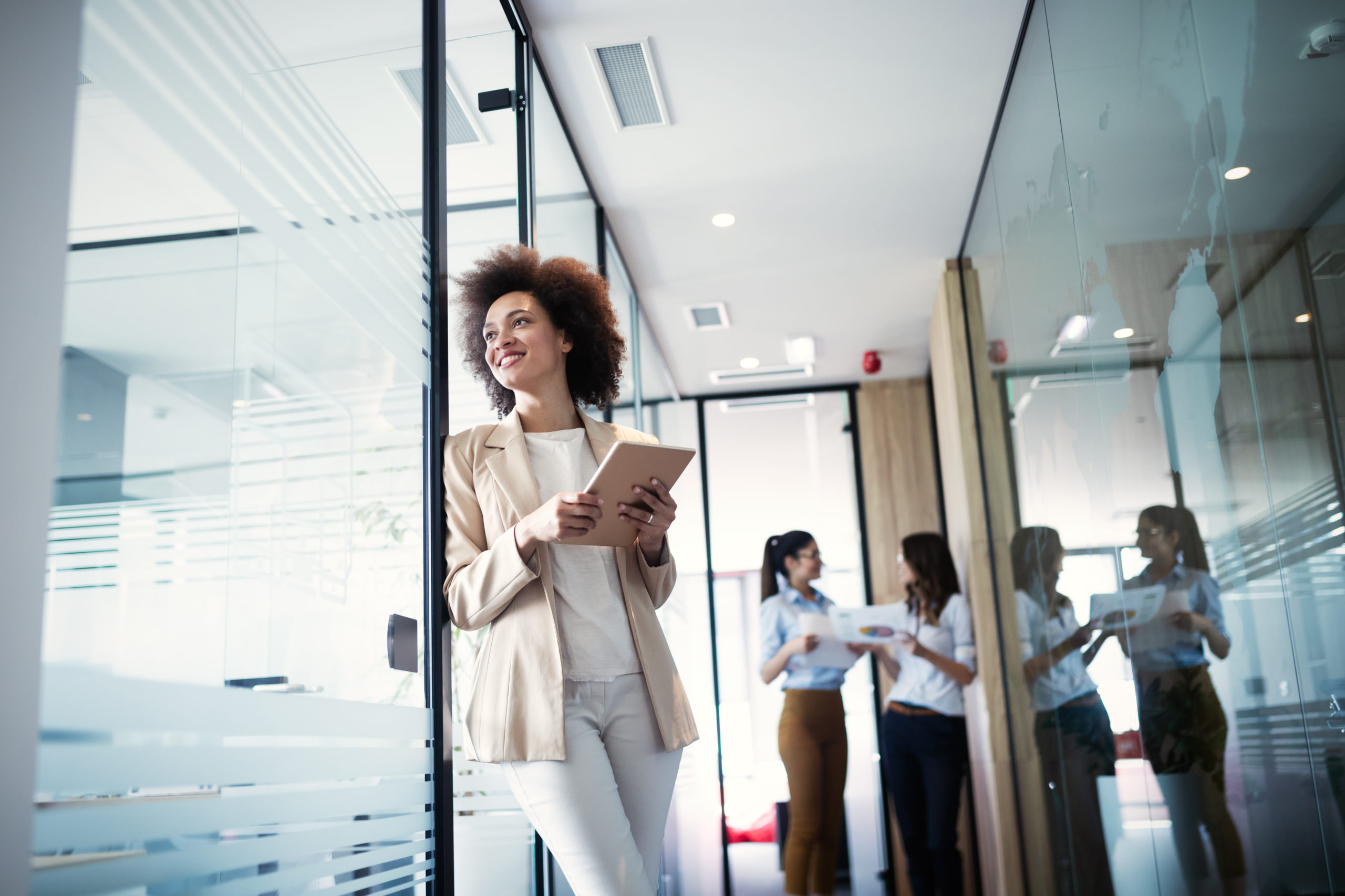 Woman of colour consultant standing in office hallway looking into the distance through glass wall and two women certification agents in the background engaged in a review conversation